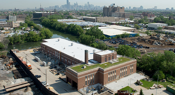 A red brick building with grass on the roof sits on the edge of a small river. A city skyline is seen in the distance.